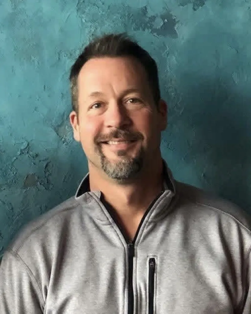 Danny Powell, smiling, in front of a teal plaster wall.